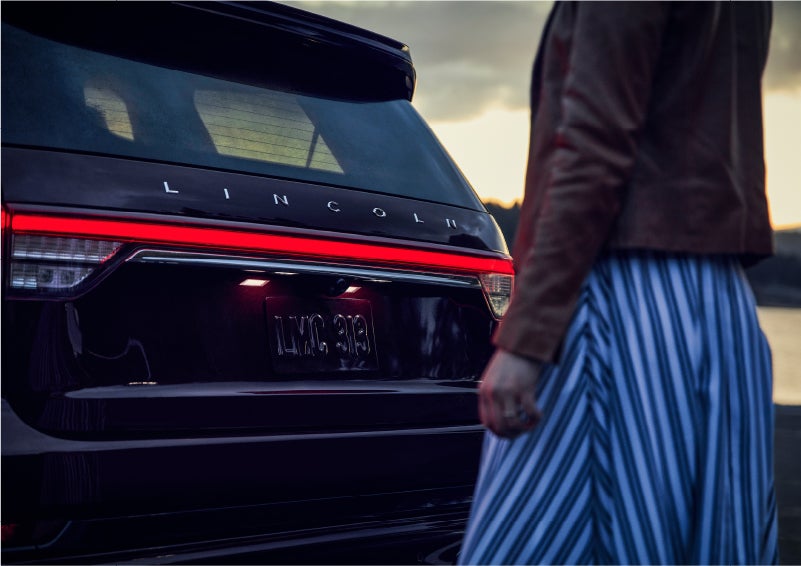 A person is shown near the rear of a 2023 Lincoln Aviator® SUV as the Lincoln Embrace illuminates the rear lights | Pilson Lincoln in Mattoon IL